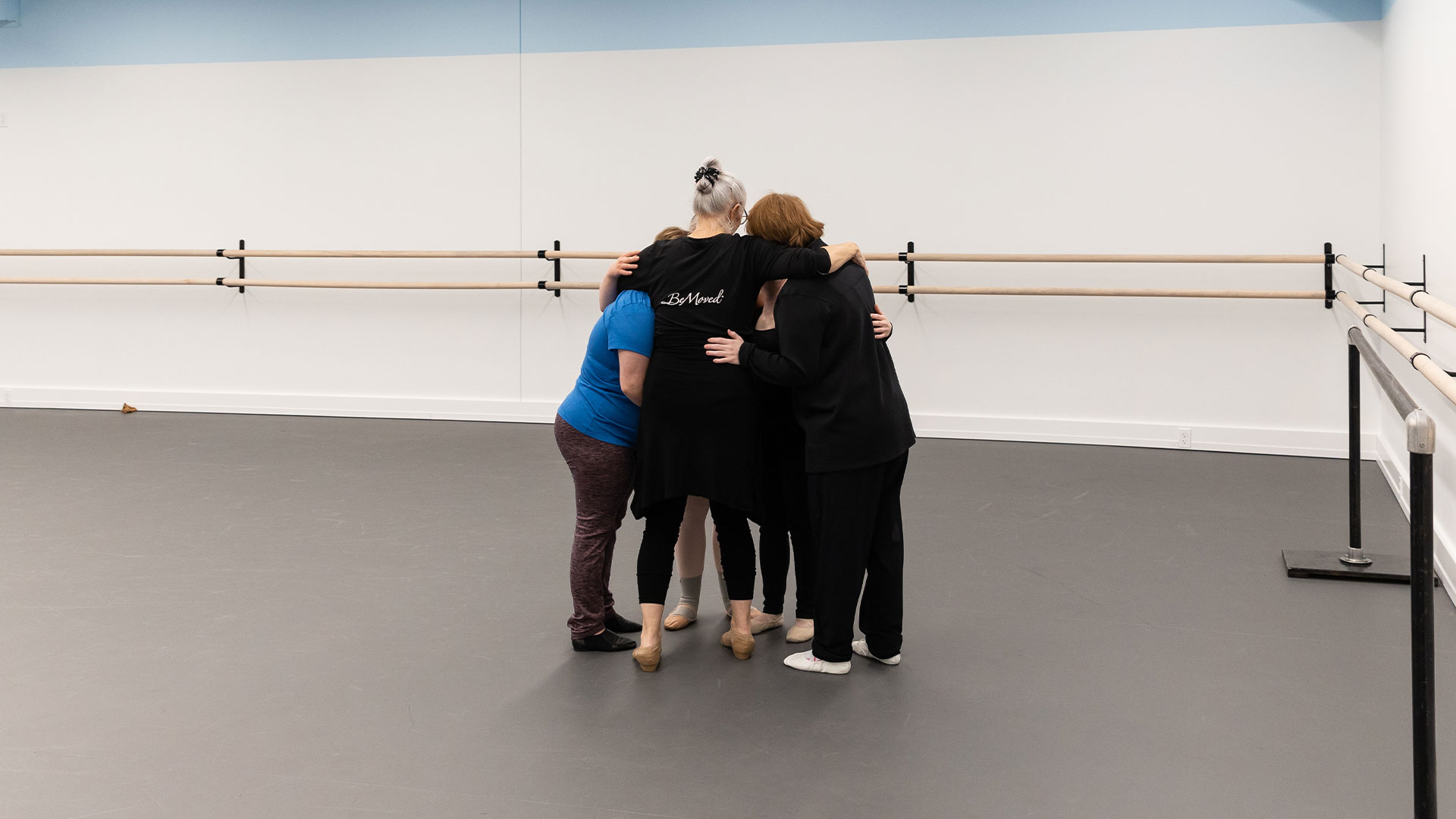 A group of adult dancers and their teacher embrace in a group hug.